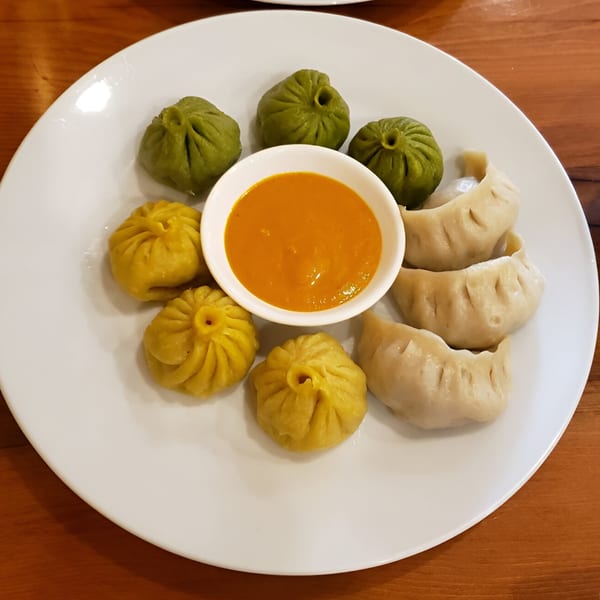 A white plate with green, yellow, and white momos surrounding a bowl of orange dipping sauce.